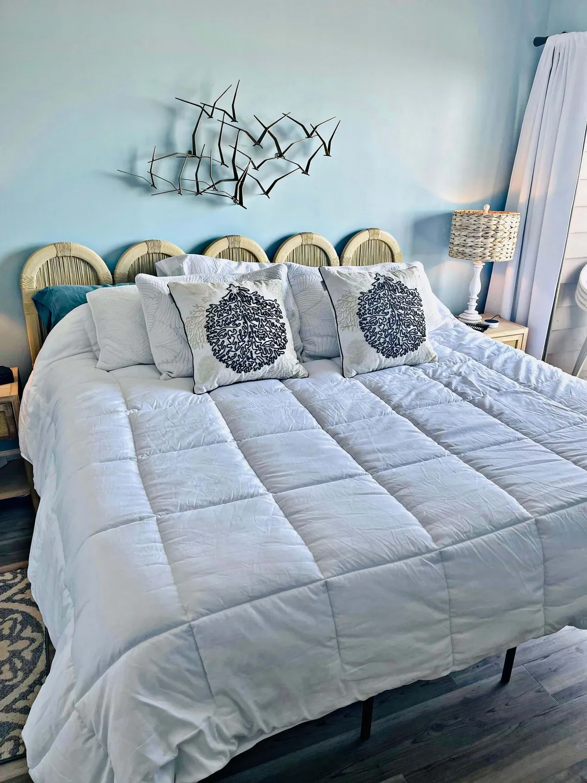 A serene bedroom featuring a plush bed dressed in a white comforter with decorative pillows, softly illuminated by natural light. Curtains partially drawn frame the view, and a tasteful art piece hangs above the headboard, creating a cozy and tranquil atmosphere ideal for relaxation.