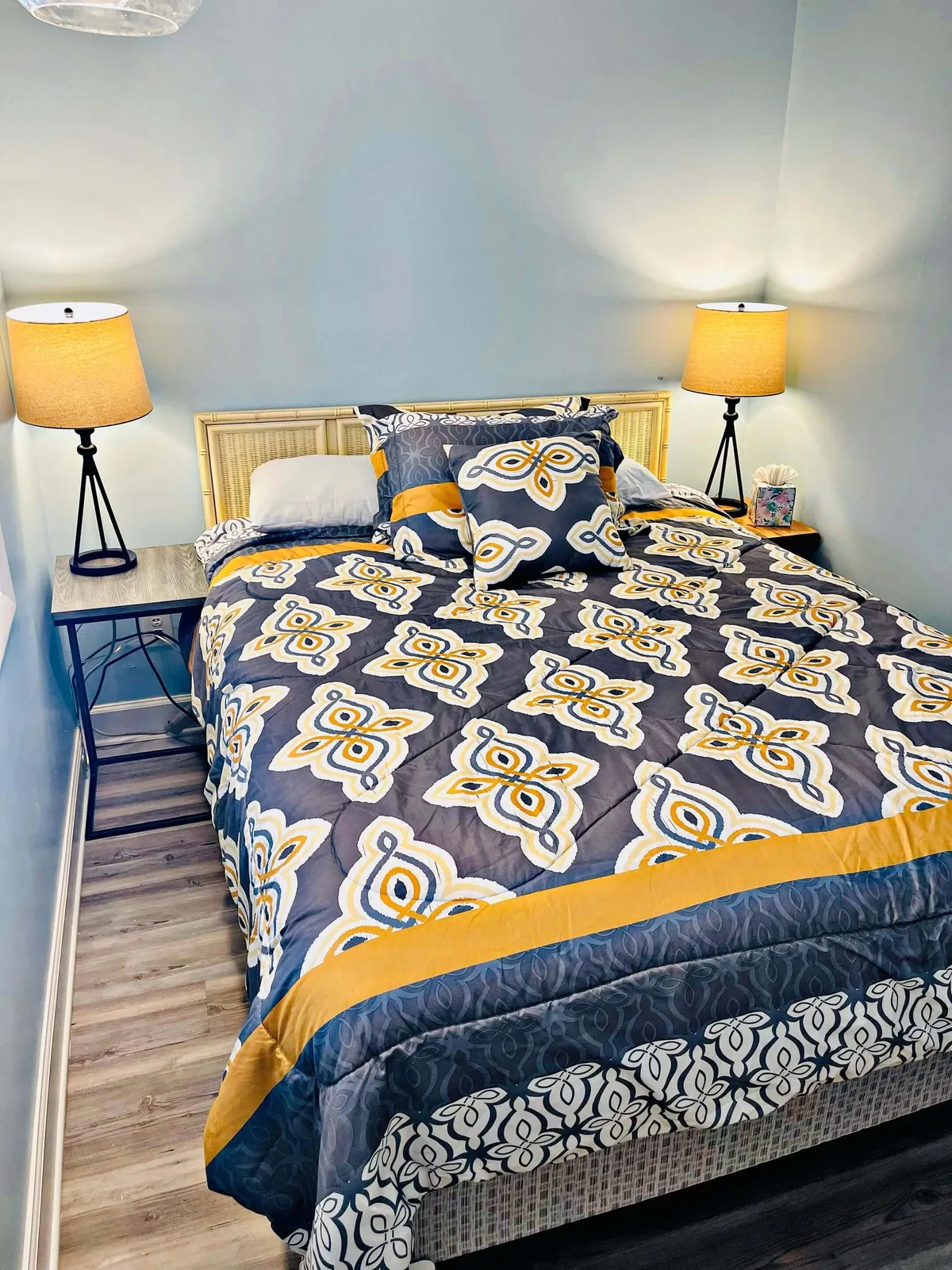 A warm, inviting bedroom with a patterned comforter on the bed, illuminated by soft lighting from bedside lamps on matching nightstands. The setting feels intimate and cozy, perfect for relaxing, reading, or unwinding after a day by the beach.