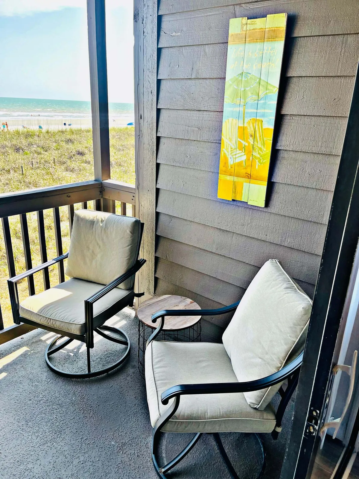 An oceanfront patio with two comfortable swivel chairs and a small wooden side table, set against a backdrop of the sparkling sea. Colorful beach-themed wall art adds a playful touch, while the space is perfect for sipping coffee or enjoying the salty breeze and sunset views.