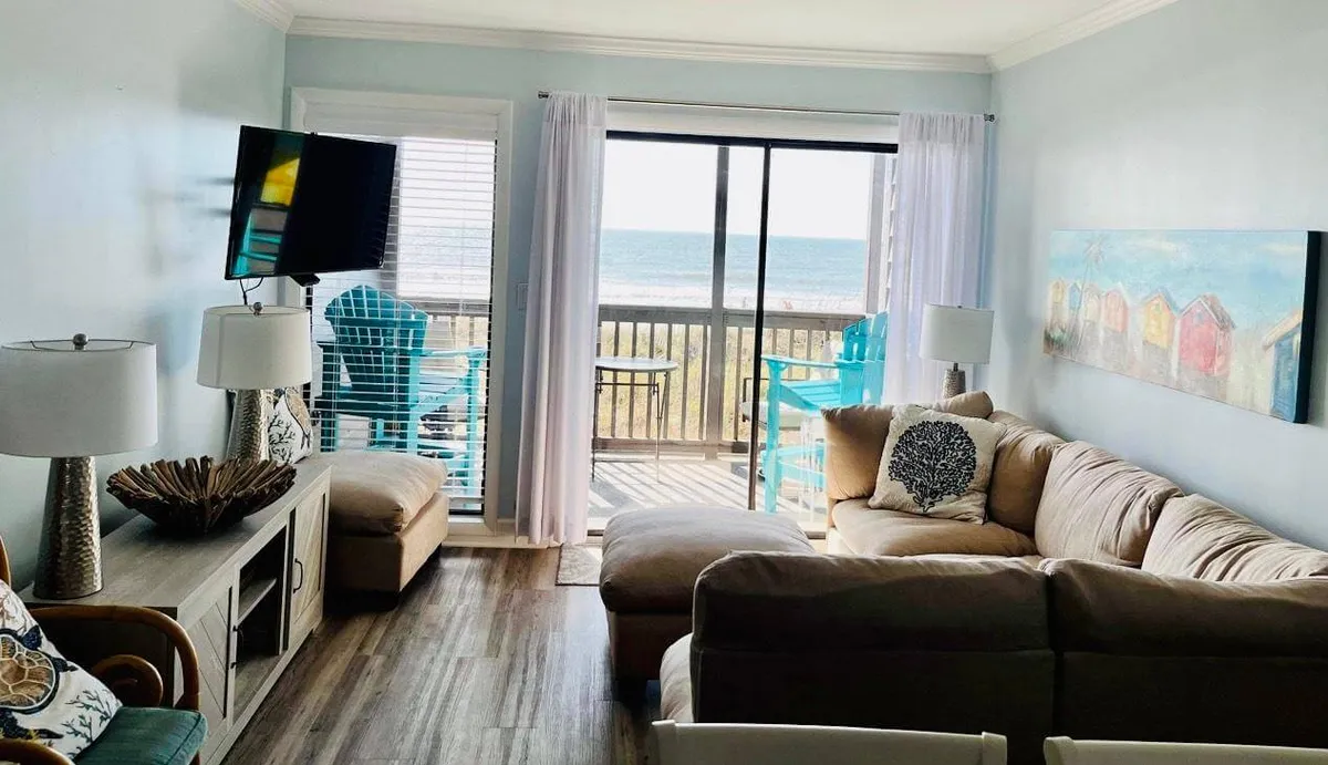 A spacious living room with a large window framing expansive ocean views. The room features a comfortable sectional sofa, colorful beach-themed artwork, and stylish lamps that create a cozy and tranquil ambiance. The setup is ideal for relaxing, socializing, or watching TV while enjoying the ocean panorama.
