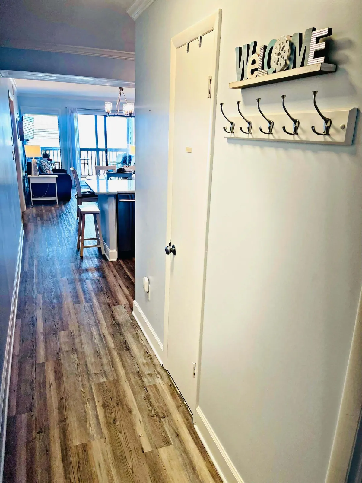 A welcoming entryway that seamlessly leads to a visible kitchen area and living space. The scene includes a 'welcome' sign and a glimpse of the kitchen, capturing the transition between spaces and the inviting atmosphere of the home.