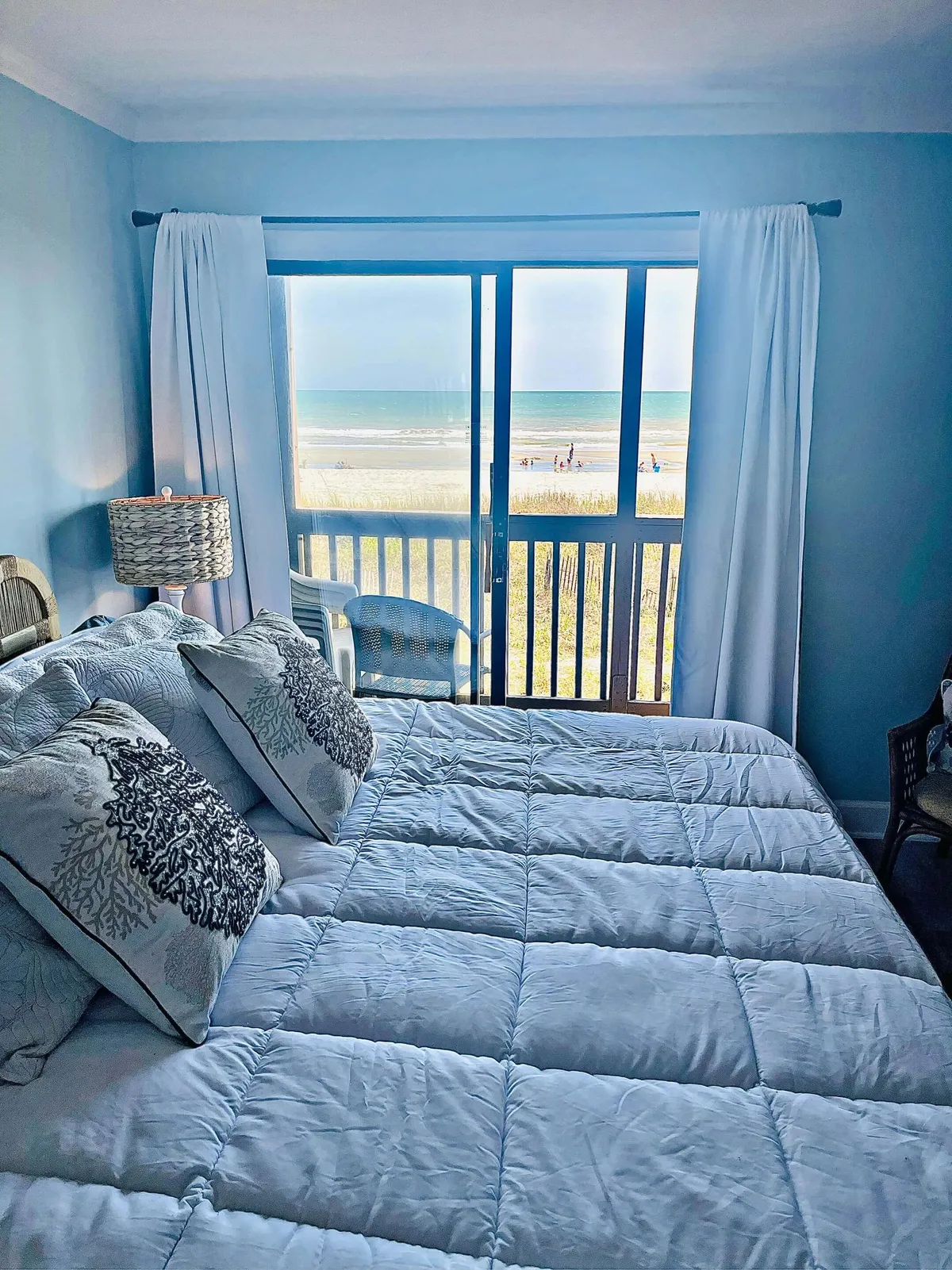 A serene bedroom with a large window offering sweeping views of the beach and ocean. Decorative pillows and a stylish lamp create an inviting atmosphere, while natural sunlight streams in, connecting the interior space directly to the beach outside.