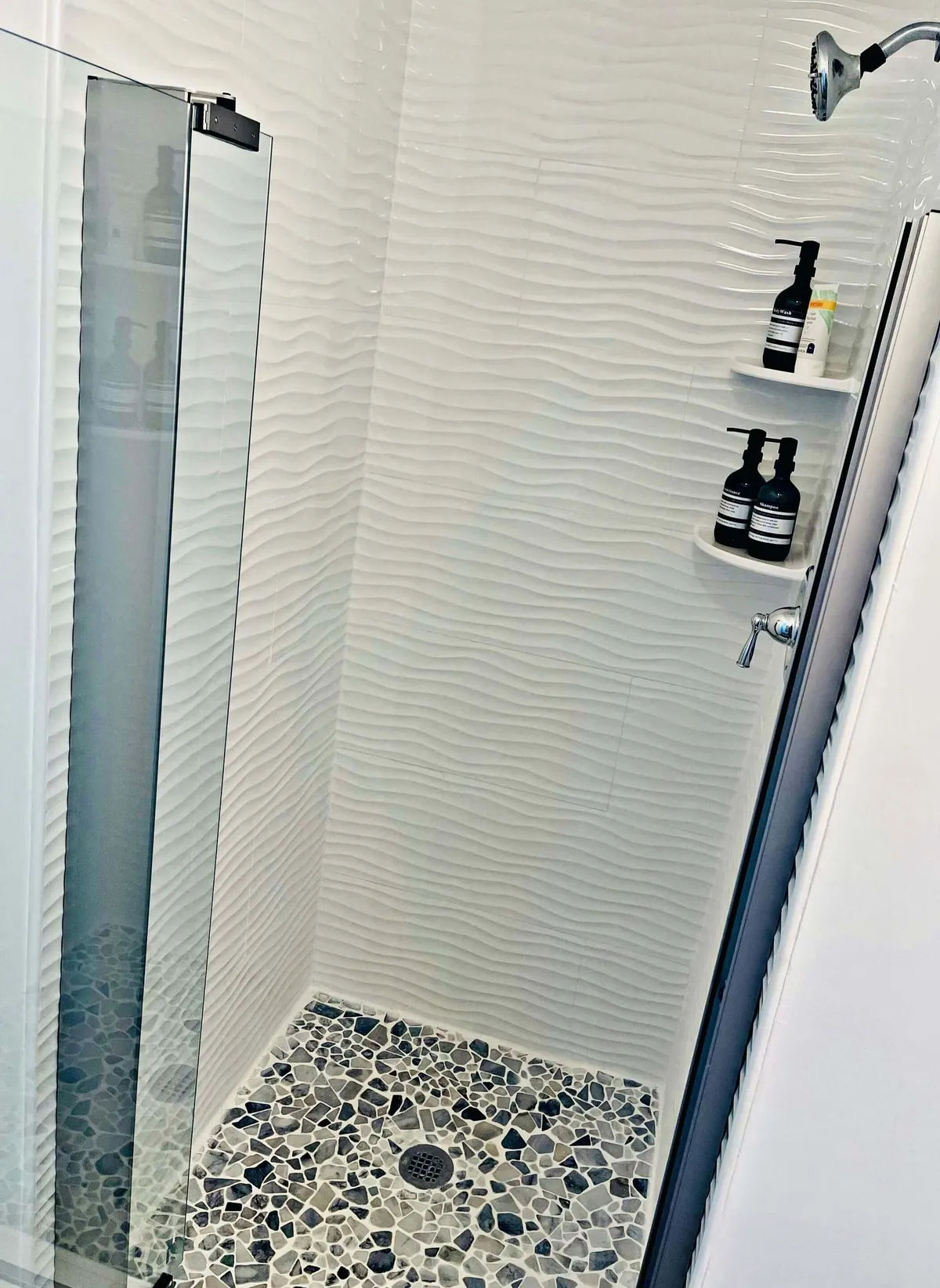 A modern ensuite bath with textured white walls, pebble stone flooring, and elegant shower fixtures. Thoughtfully placed toiletries and soft lighting create a spa-like, rejuvenating atmosphere.