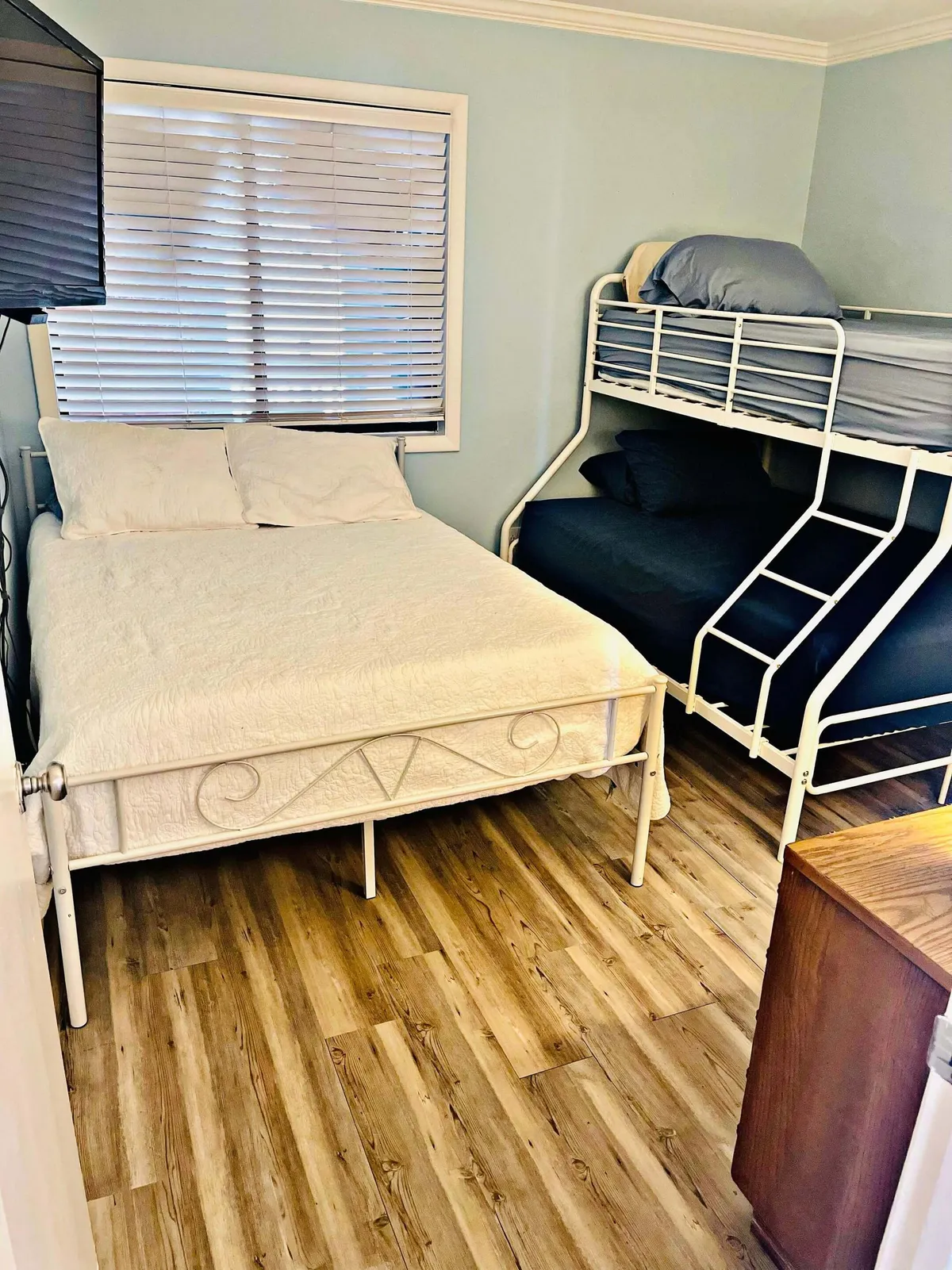 A bedroom designed for versatility features a bunk bed and a double bed, with a window letting in soft daylight. There's a television for entertainment and plenty of space for families or groups.