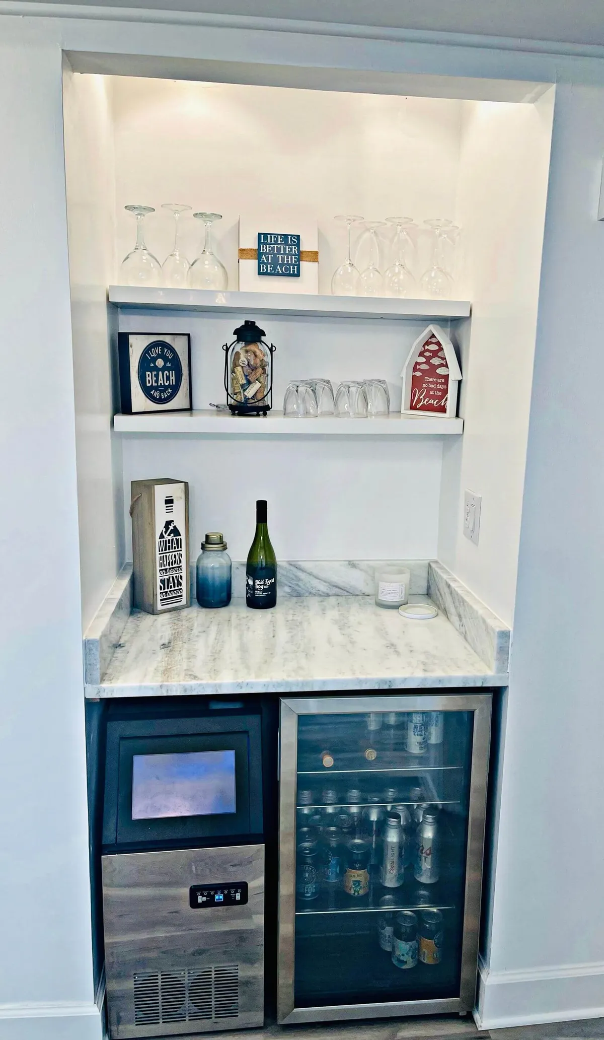 A cheerful, laid-back space featuring a set of shelves with glassware, decorative beach-themed signs, and a beverage cooler. The area is designed for entertaining and casual gatherings, with hints of sun and relaxation from the nearby beach visible through the décor.